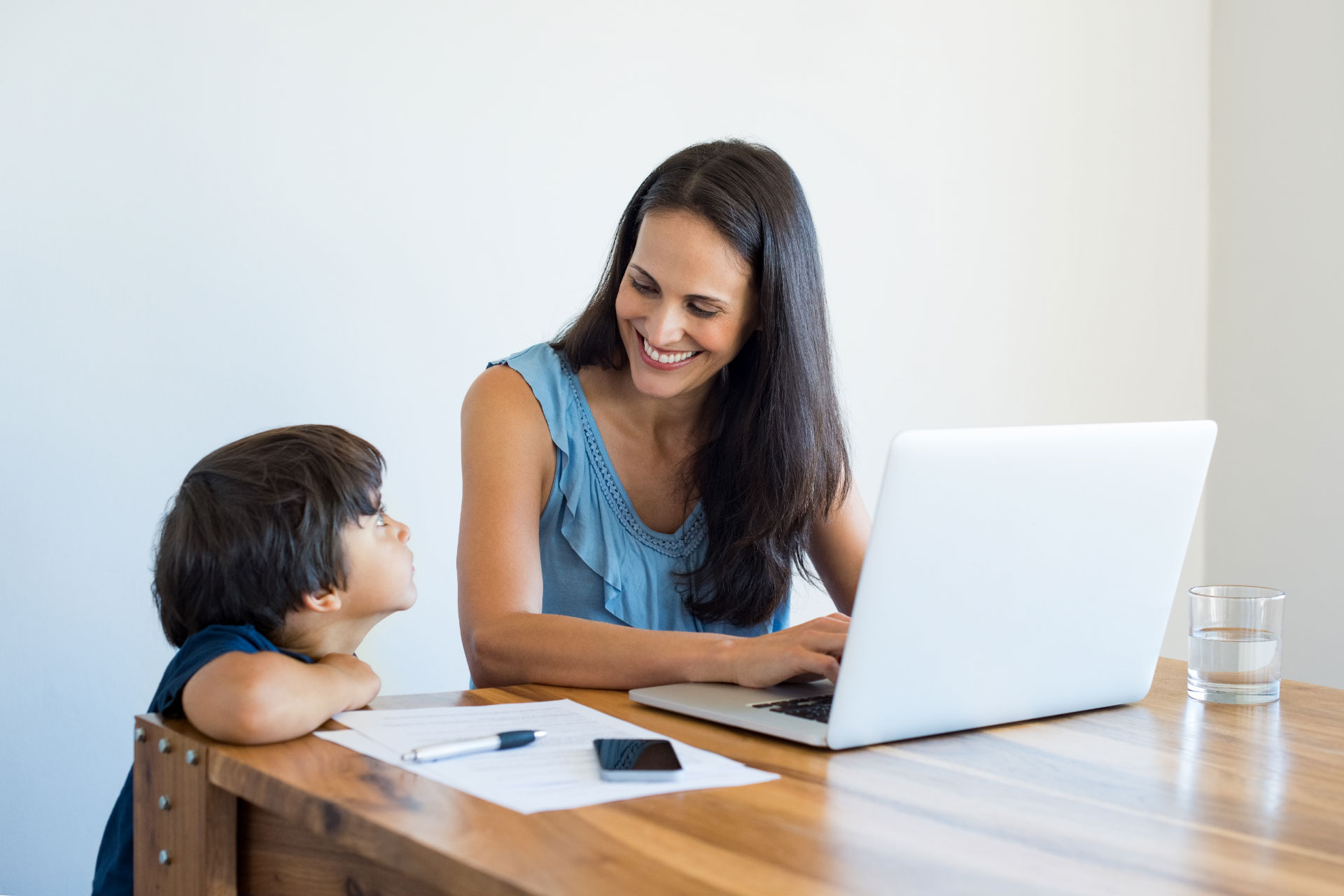 Smiling and confident mother is working at home with her laptop on a dining table, while speaking to her son who's standing beside her and looking up at her.